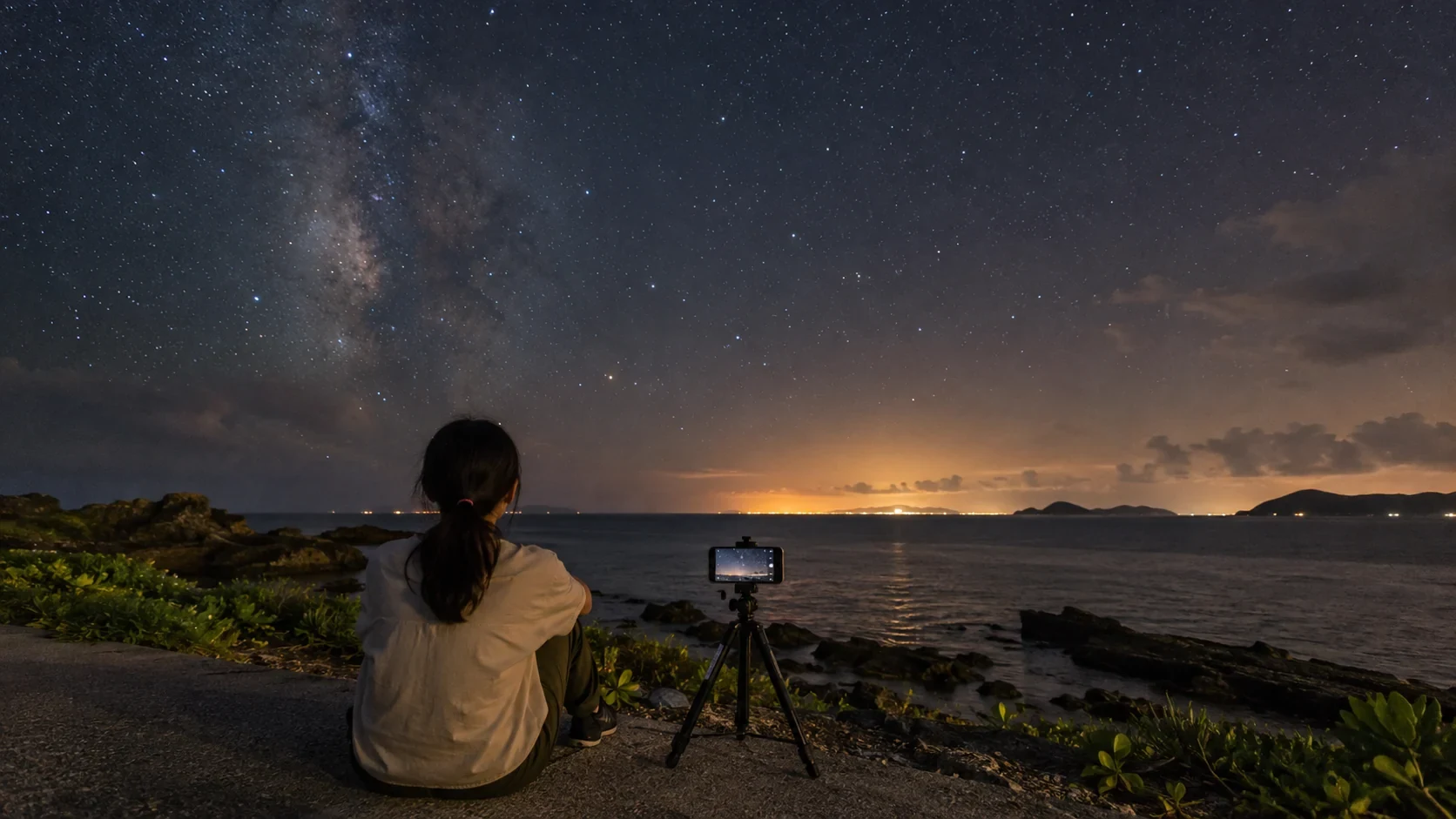 古宇利島の海辺でスマートフォンを三脚に固定し、星空を見上げる旅人の写真調イメージ